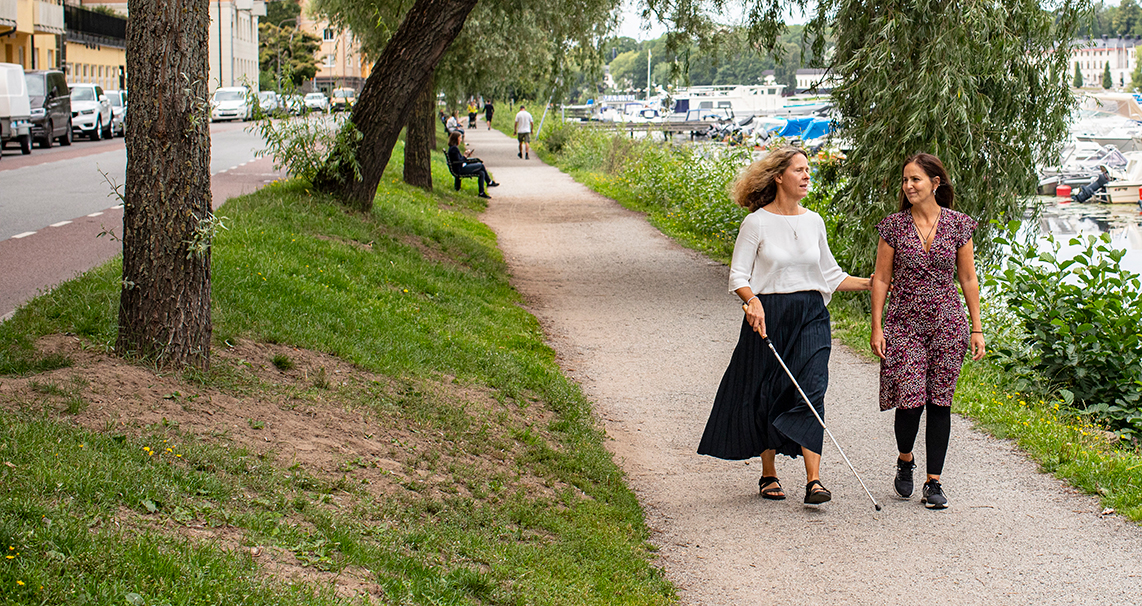 Två kvinnor promenerar tillsammans på ett promenadstråk vid vattnet. Den ena har vit käpp och blir ledsagad av den andra.