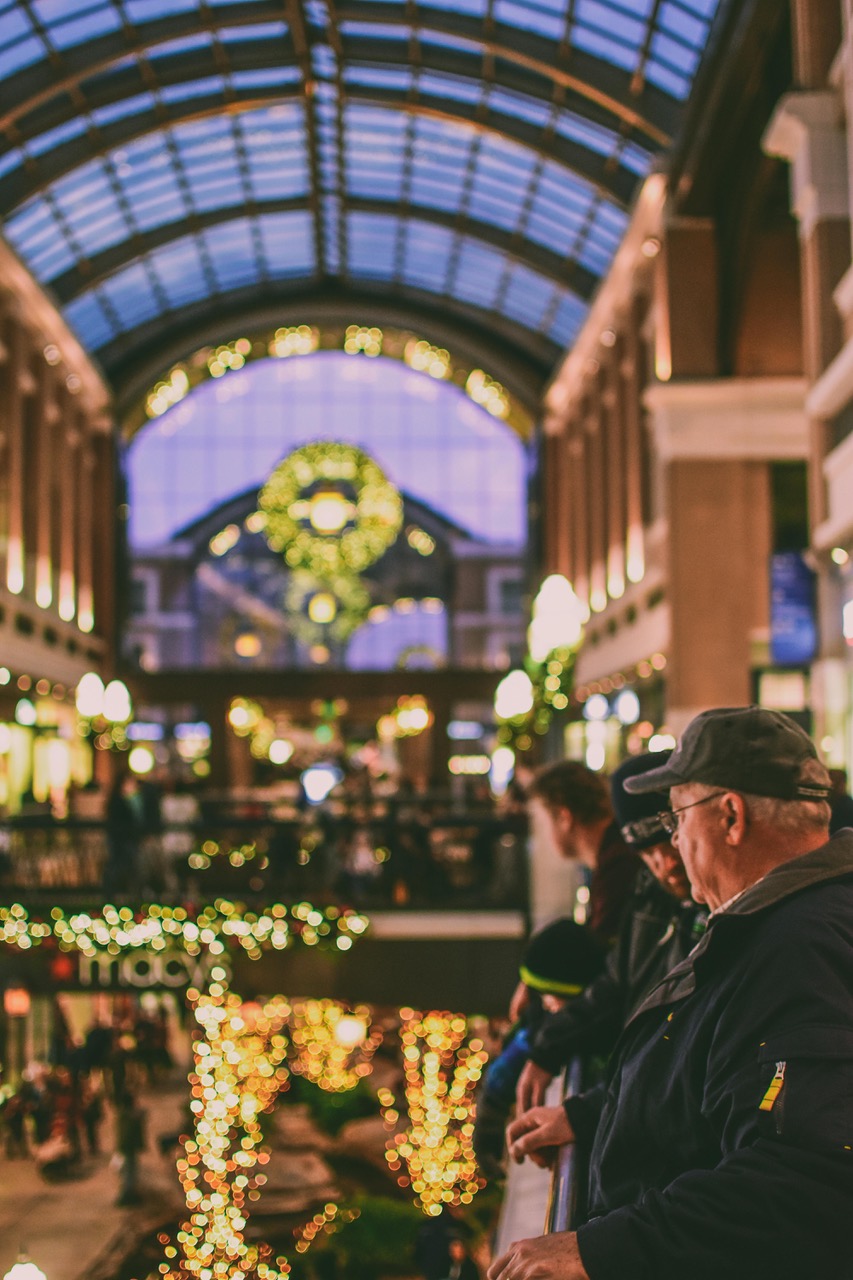 En rymlig hall, det kan vara ett köpcentrum, är julpyntad med ljusslingor och en stor krans i fönstret. I förgrunden syns en man med keps och glasögon. 