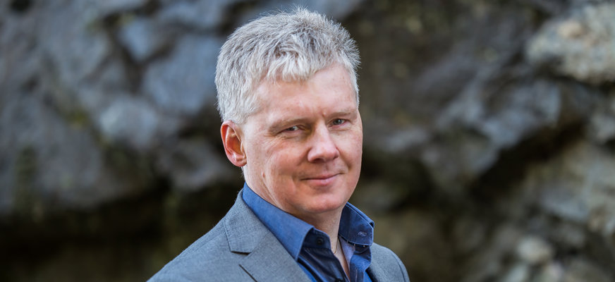 Niklas Mattsson i grå kostym, blå skjorta framför bergvägg.