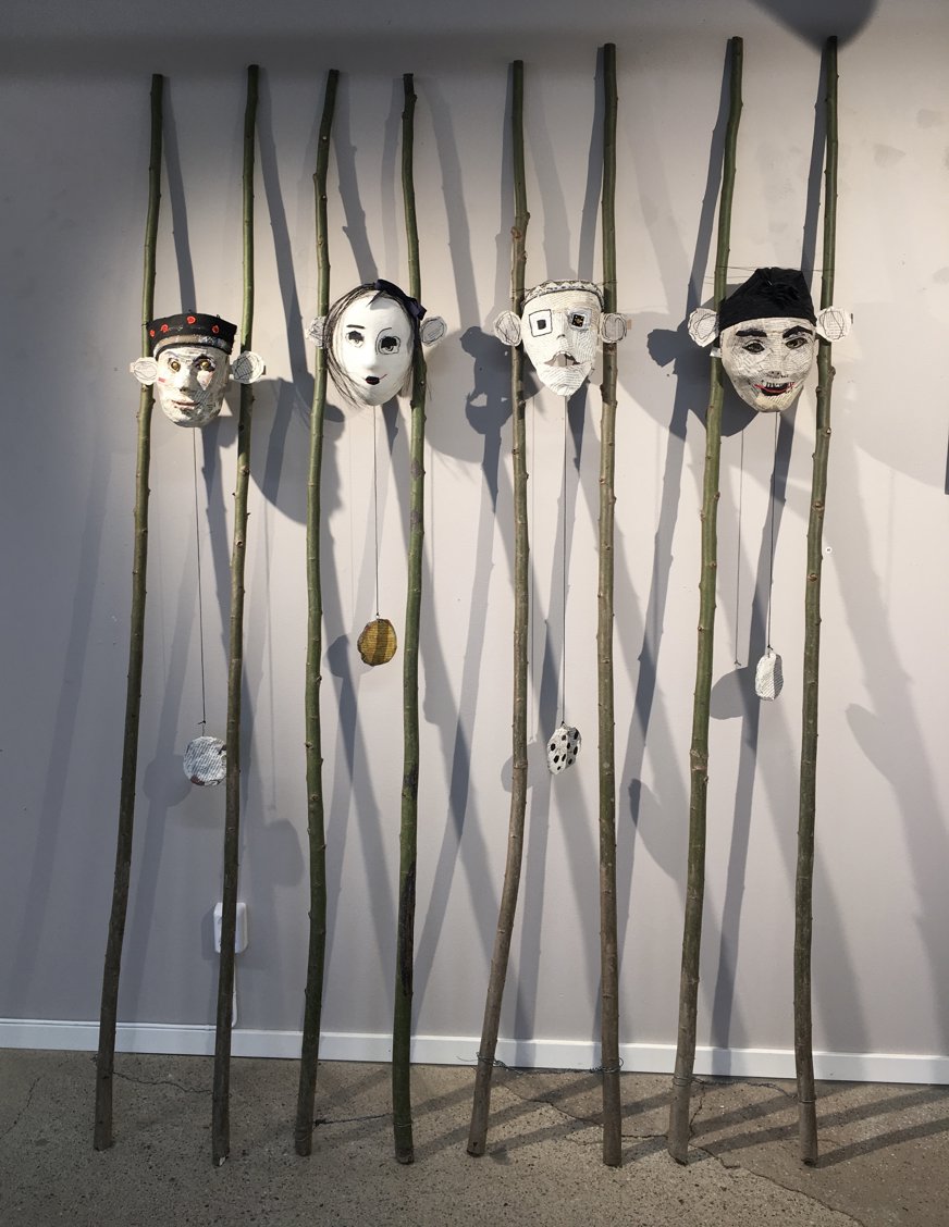 Det här konstverket består av fem masker som är upphängda på långa, tunna stavar av bambu eller liknande naturligt material. Varje mask har en unik ansiktsdesign och uttryck, och visar en variation av stilar och känslor.  Från vänster till höger:  Den första masken har ett ansikte målat i jordnära toner med runda, vidöppna ögon och ett svart pannband dekorerat med röda cirklar. Den andra masken är målad i vitt, med mörka ögon och ett förvånat uttryck, som om den blivit tagen på sängen. Den tredje masken har ett rektangulärt ansikte med mustasch och ett ögonlapp eller monokel på ena sidan, och ett litet, allvarligt uttryck. Den fjärde masken har en mer abstrakt design med asymmetriska ögon och detaljerad skuggning, vilket ger den ett lite udda utseende. Den femte masken har ett leende uttryck, med överdrivet röda läppar och en mörk mössa, vilket ger den ett glatt intryck. Nedanför varje mask hänger små, texturerade föremål från snören fästa på stavarna, vilket ger en extra dimension till verket. Skuggorna som maskerna och stavarna kastar på väggen skapar ett djup i bilden. Konstverket har en jordnära, organisk känsla och kan antyda teman som individualitet, kulturell identitet eller en representation av olika personligheter.