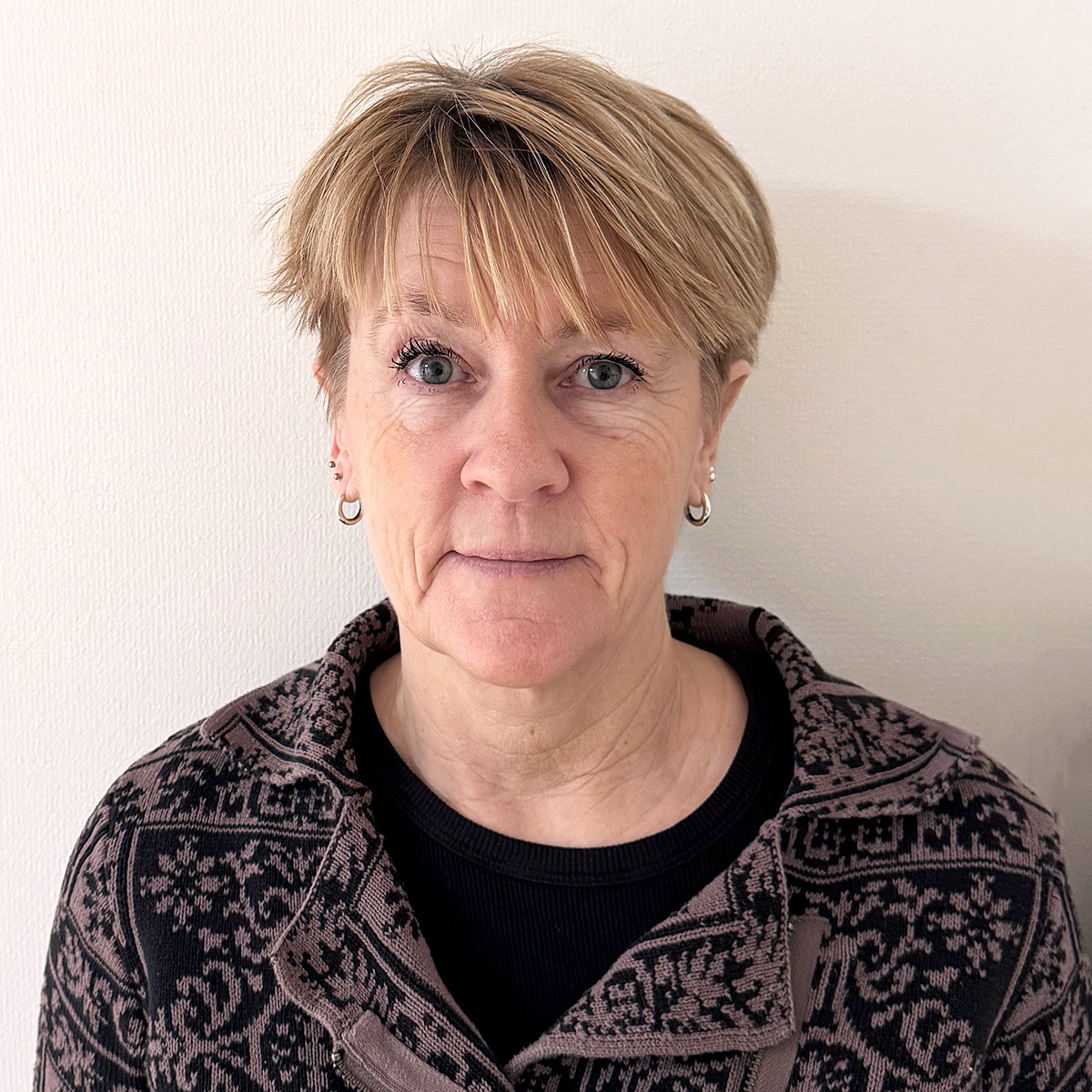 Porträttbild på Jeanet. Kvinna i 60-årsåldern med kort, mellanblont hår med lugg. Jeanet har blå ögon och ser med alert blick in i kameran. Hon bär en stickad tröja mönstrad i lila och svart. I öronen har hon flera ringar i silver.