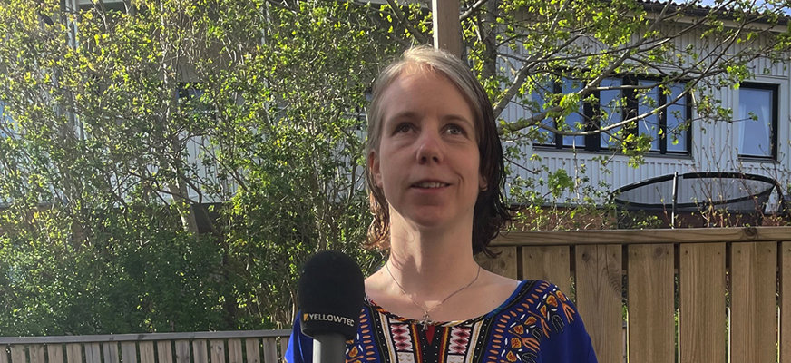 Isabelle står utomhus och håller leende upp en mikrofon framför sig. Hon är iförd en mörkblå kortärmad klänning med mönstrat bröst i vitt och orange.