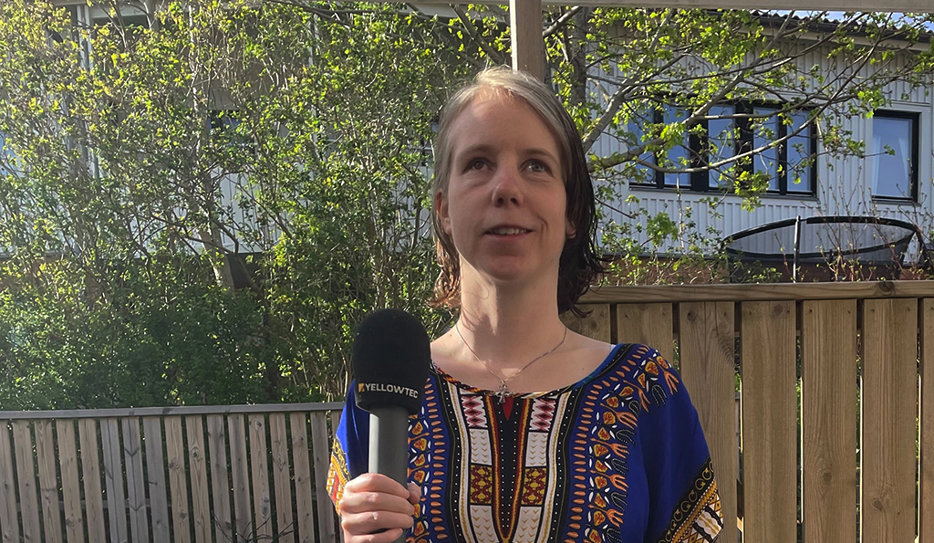 Isabelle Ulfsdotter. Kvinna i kort frisyr som står utomhus och håller leende upp en mikrofon framför sig. På sig har hon en blå, kortärmad klänning med mönstrat bröst.