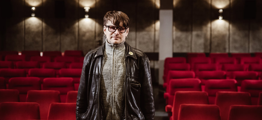 Finn Hellman står i en biosalong med tomma stolar i bakgrunden. Han har glasögon och skinnjacka.