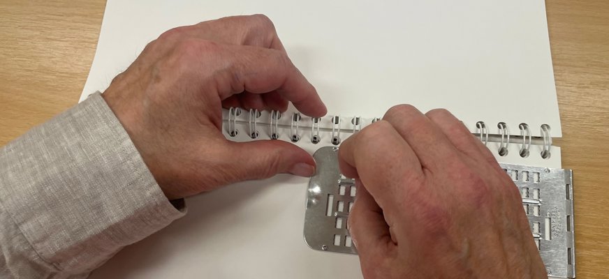 Händer som skriver punktskrift för hand med reglett och syl.