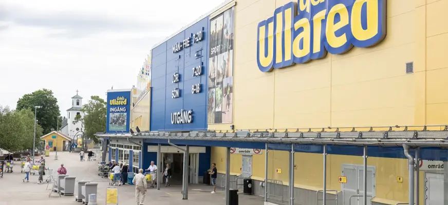 Bilden visar Entrén på varuhuset Gekås i Ullared. Byggnaden är gul med blå detaljer vilket är Gekås färger. Vädret är halvklart och det ser ut att vara sommar.