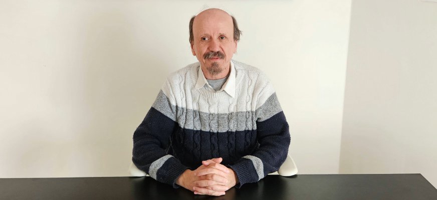 En man sitter vid ett svart bord med händerna knäppta framför sig. Han har kort, tunnare hår upptill och mustasch samt bär en stickad tröja i vitt, grått och mörkblått över en ljus skjorta. Bakgrunden är en ljus vägg och bilden är tagen rakt framifrån i ögonhöjd.