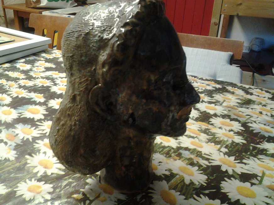 Ett skulpterat människohuvud i profil, riktat åt höger, står på ett bord med en färgglad duk med prästkragar. Huvudet är gjort i ett mörkt, grovt material som framhäver detaljer som öron, näsa, läppar och hårstruktur. Kontrasten mellan den mörka bysten och den ljusa, blommiga duken skapar en oväntad visuell effekt. I bakgrunden syns trästolar och vardagsföremål, vilket ger en hemtrevlig inramning till det konstnärliga objektet.