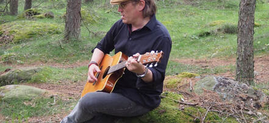 State Lycke sitter på en sten i en skogsmiljö och spelar akustisk gitarr. Han bär keps, mörk tröja och jeans, och omges av träd och grönska.