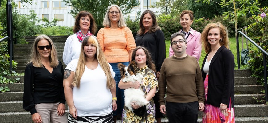 Gruppbild på kanslipersonalen stående vid stentrappa med grönskande buskar och träd i bakgrunden. Bakre raden: Helene, Lena, Jenny och Emira. Främre raden: Åsa, Linda, Carin (med hunden Harry i famnen), Alireza och Malin.