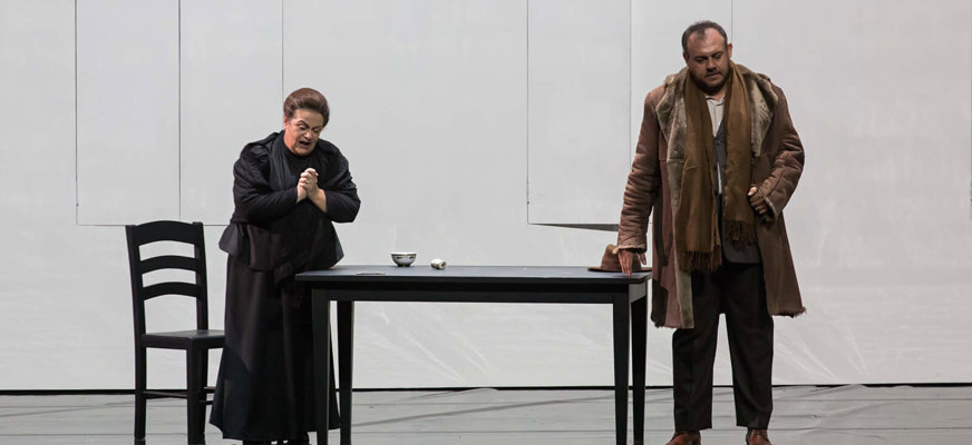 Bilden visar en man och en kvinna i en väldigt avskalad miljö. Ljus bakgrund. Kvinnan är svartklädd och mannen har mörka byxor och brun halsduk och jacka. Båda har bekymrade bistra miner och tittar åt varsitt håll